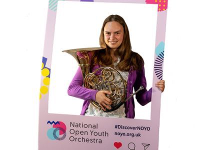 A girl with shoulder length brown hair, wearing a white top and purple cardigan holds a circular trumpet. She stands in between a customised National Open Youth Orchestra Instagram frame. She has a smile across her face.
